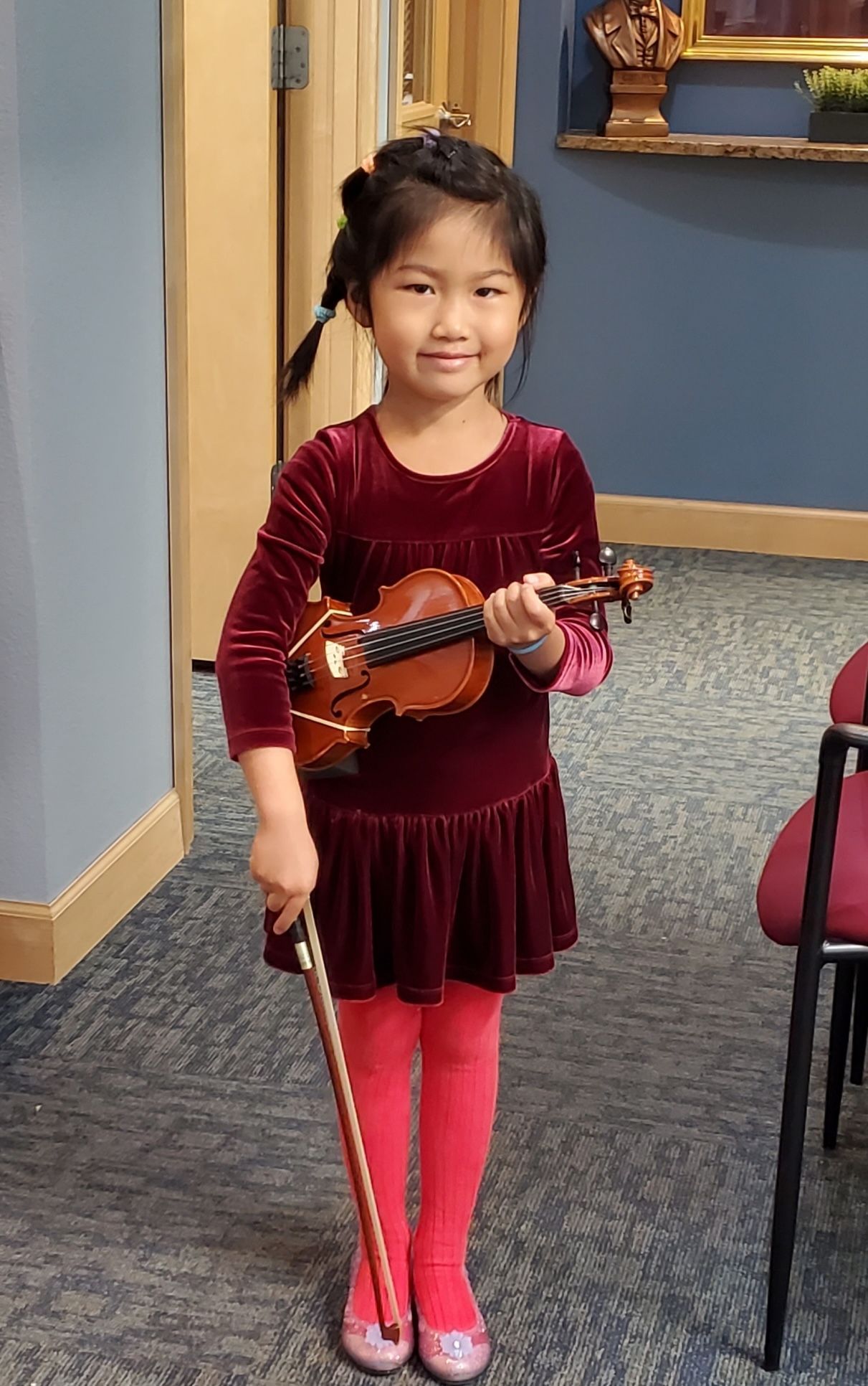 Blonde woman in a light pink dress smiles beside a piano in a wood-paneled room.