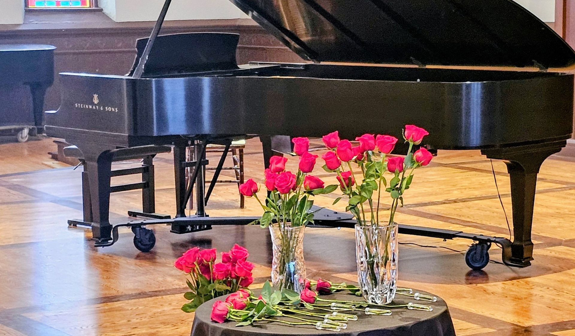 Man helps another play a piano. One wears a red shirt and plays; the other, a suit, stands next to him.