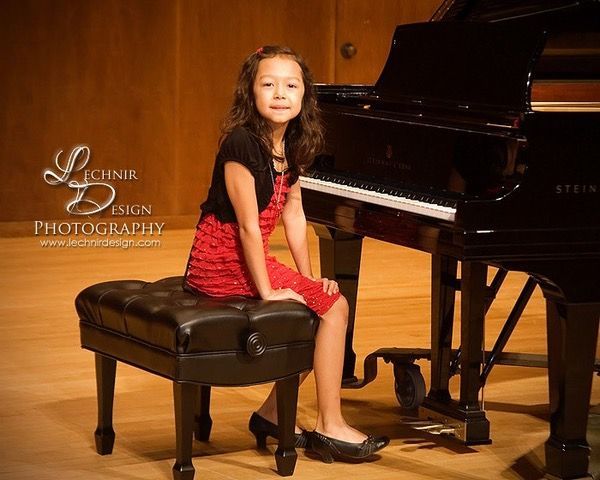 Blonde person playing violin; dark background. They wear a black shirt with red floral embroidery.