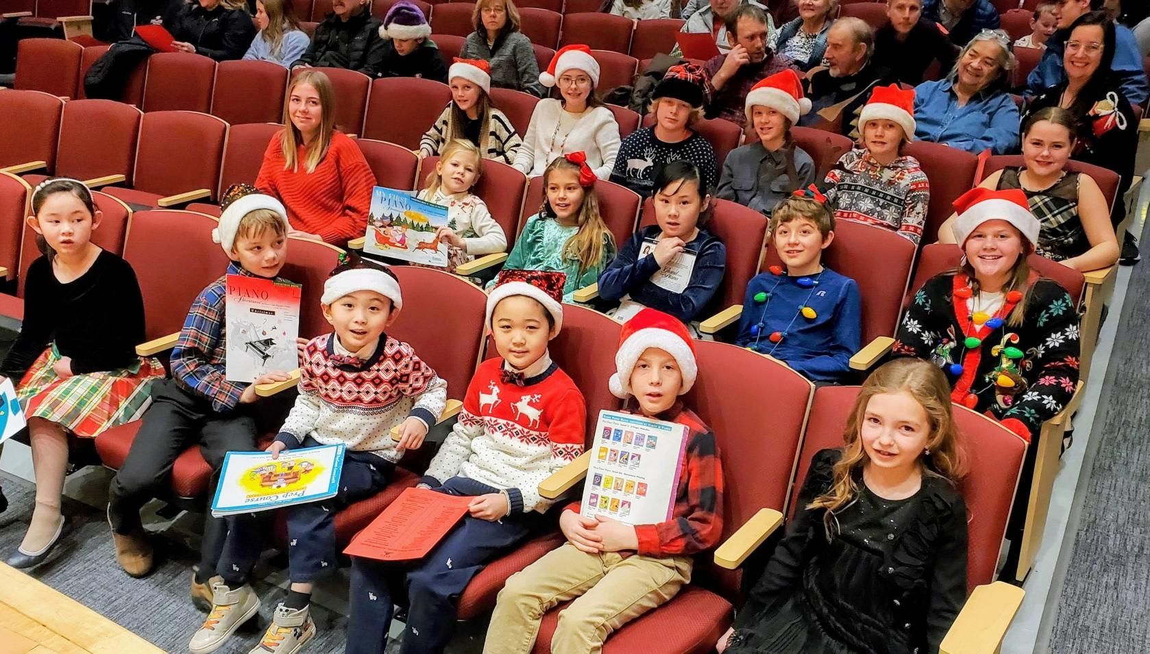 Children in Santa hats seated in theater seats, holding books, with an audience in the background.