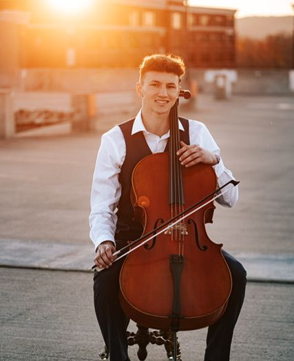Man helps another play a piano. One wears a red shirt and plays; the other, a suit, stands next to him.