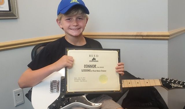 Boy holding a guitar and a certificate, smiling. He wears a blue hat and black shirt, in a room.