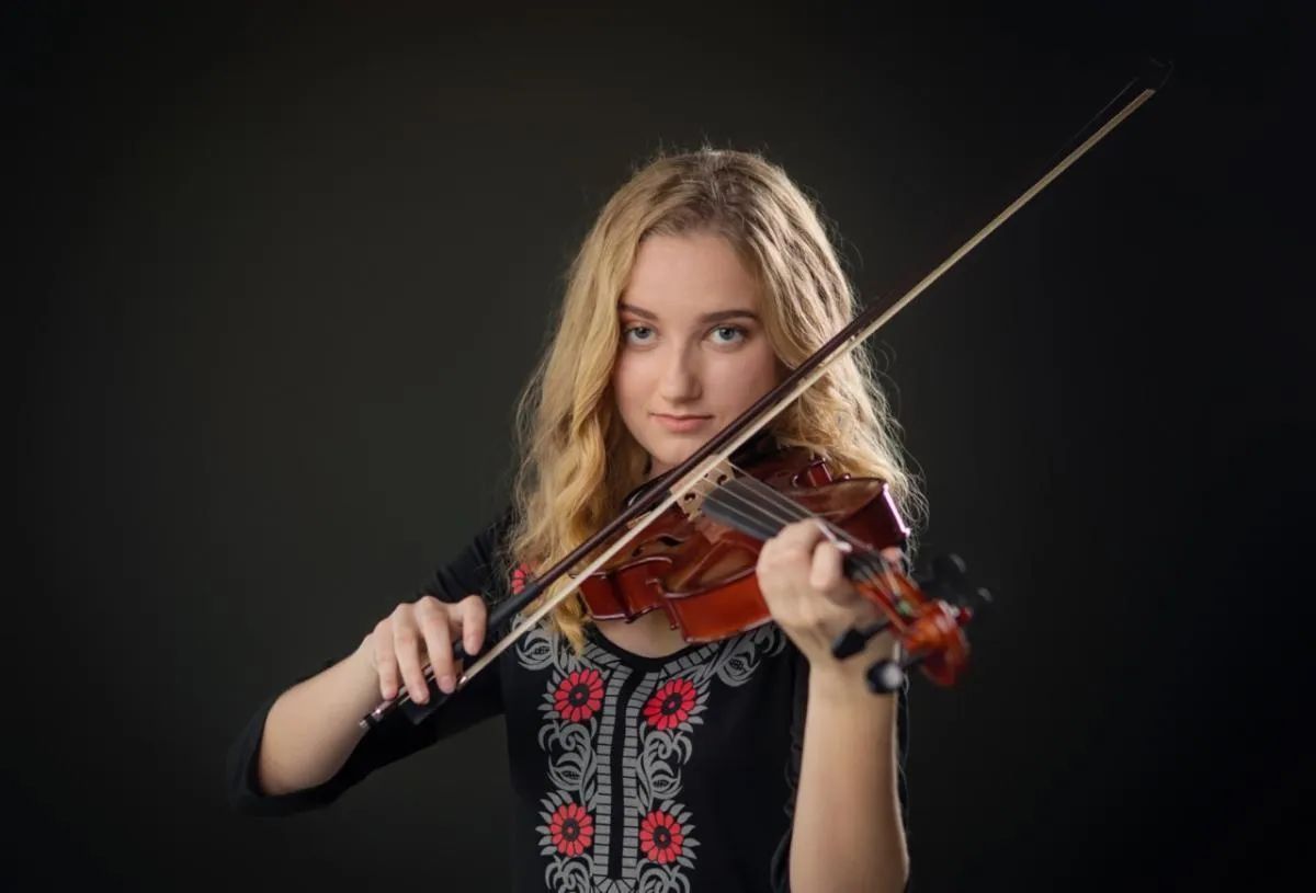 Blonde person playing violin; dark background. They wear a black shirt with red floral embroidery.
