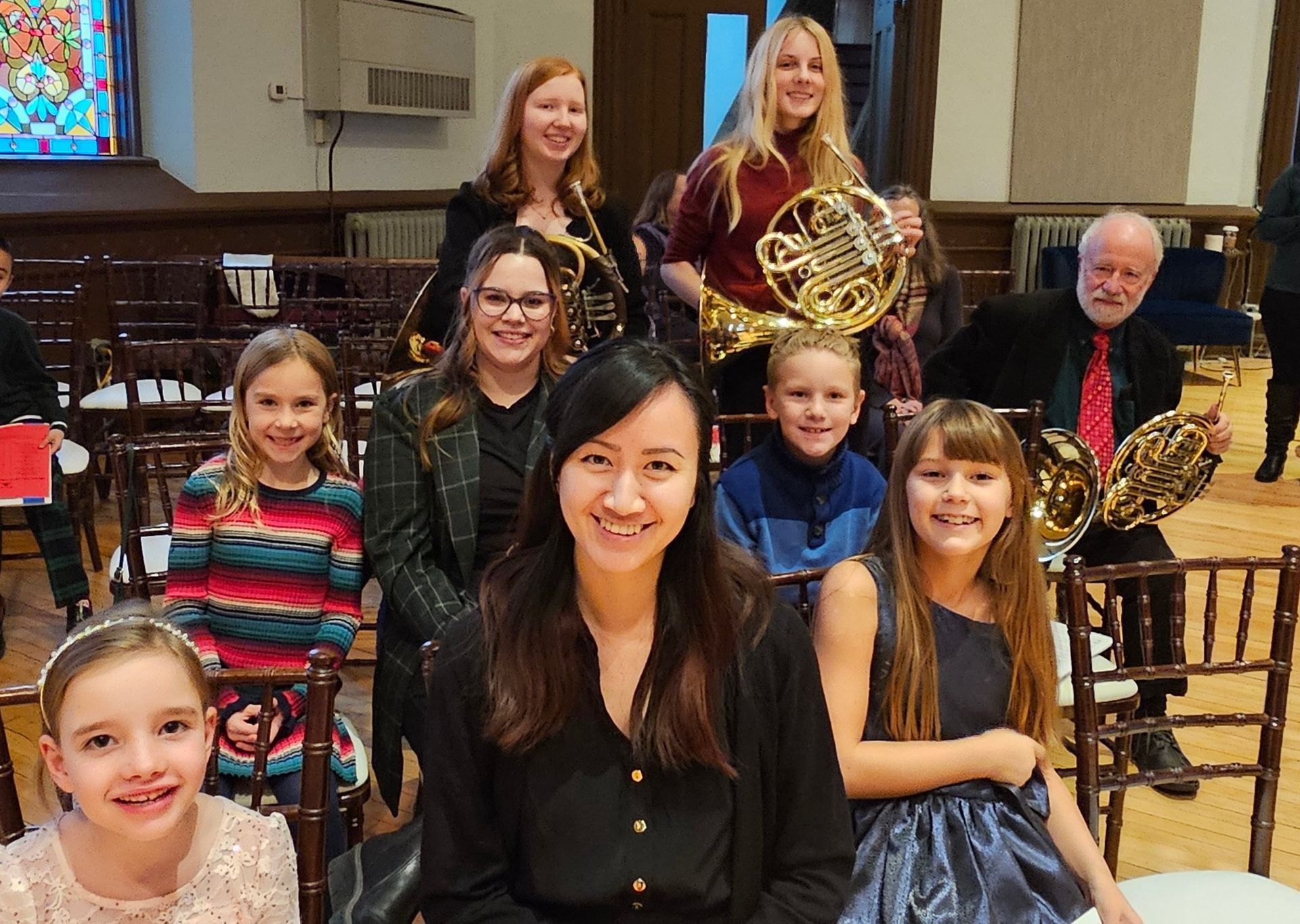 A group of people with french horns in a room with chairs. Some are smiling at the camera.