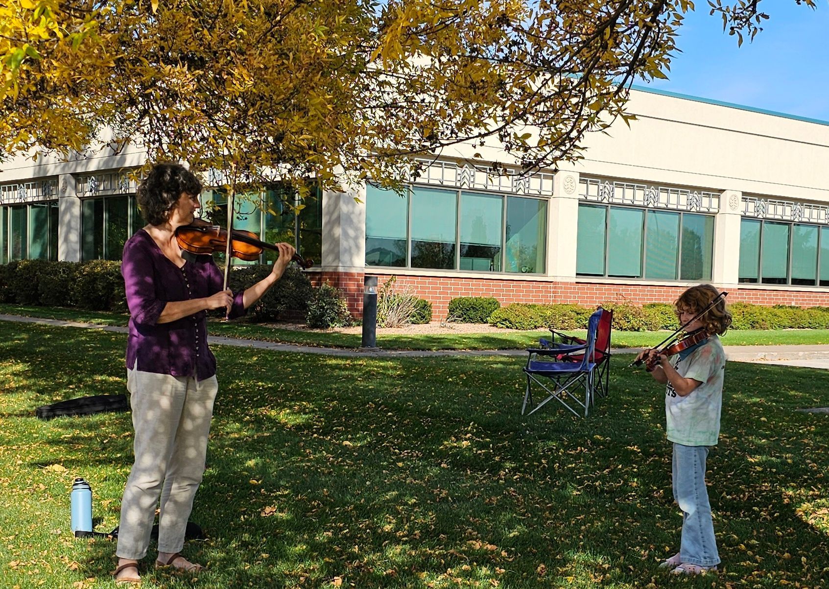 Woman and child playing violins outdoors in front of a building; autumn setting.