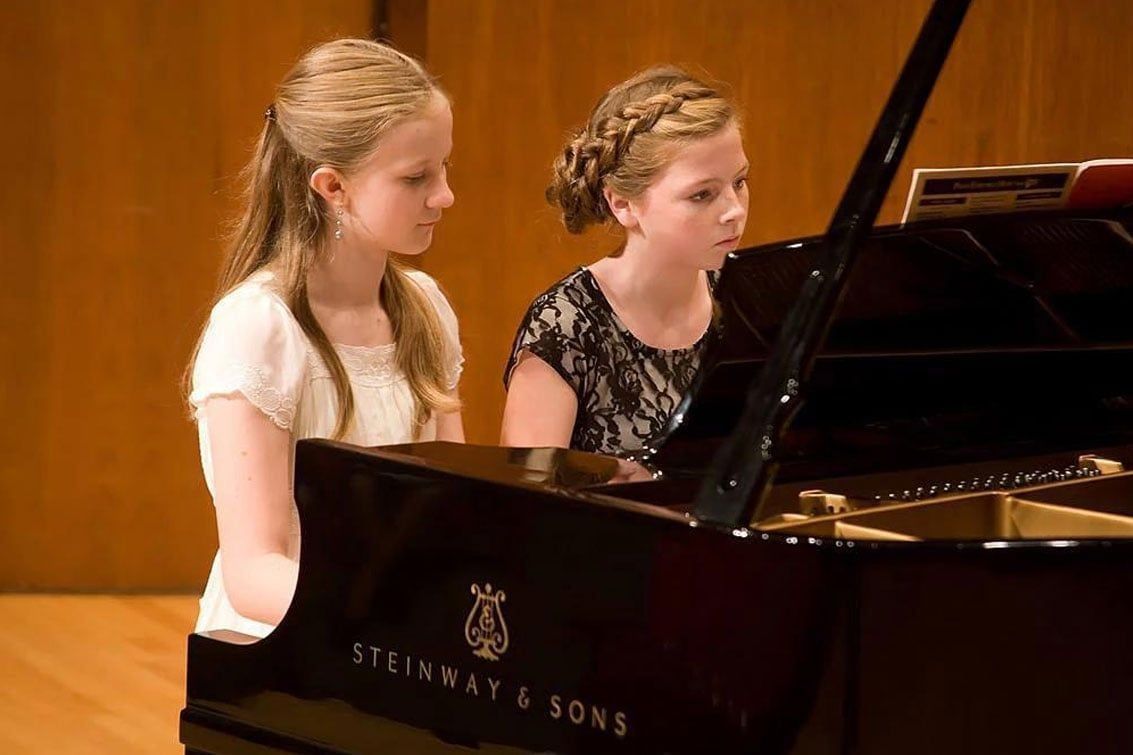 Two young girls playing a black grand piano. One wears white, the other black. Wooden background.