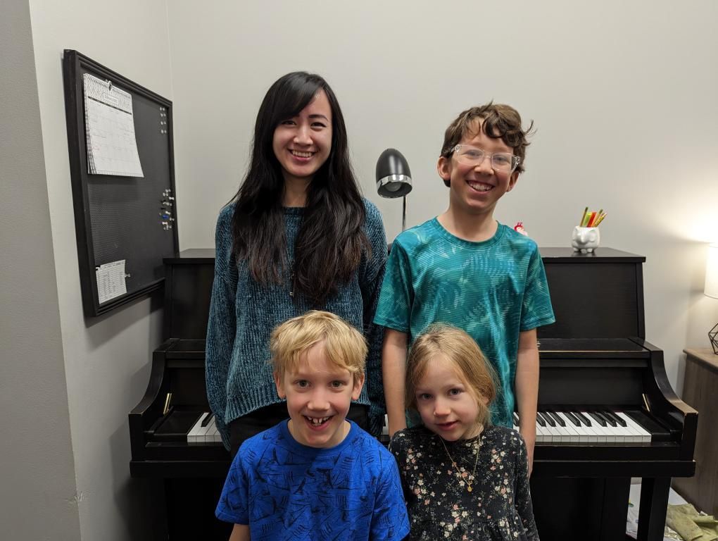 Woman and three children smiling in front of a piano in a room with a bulletin board.