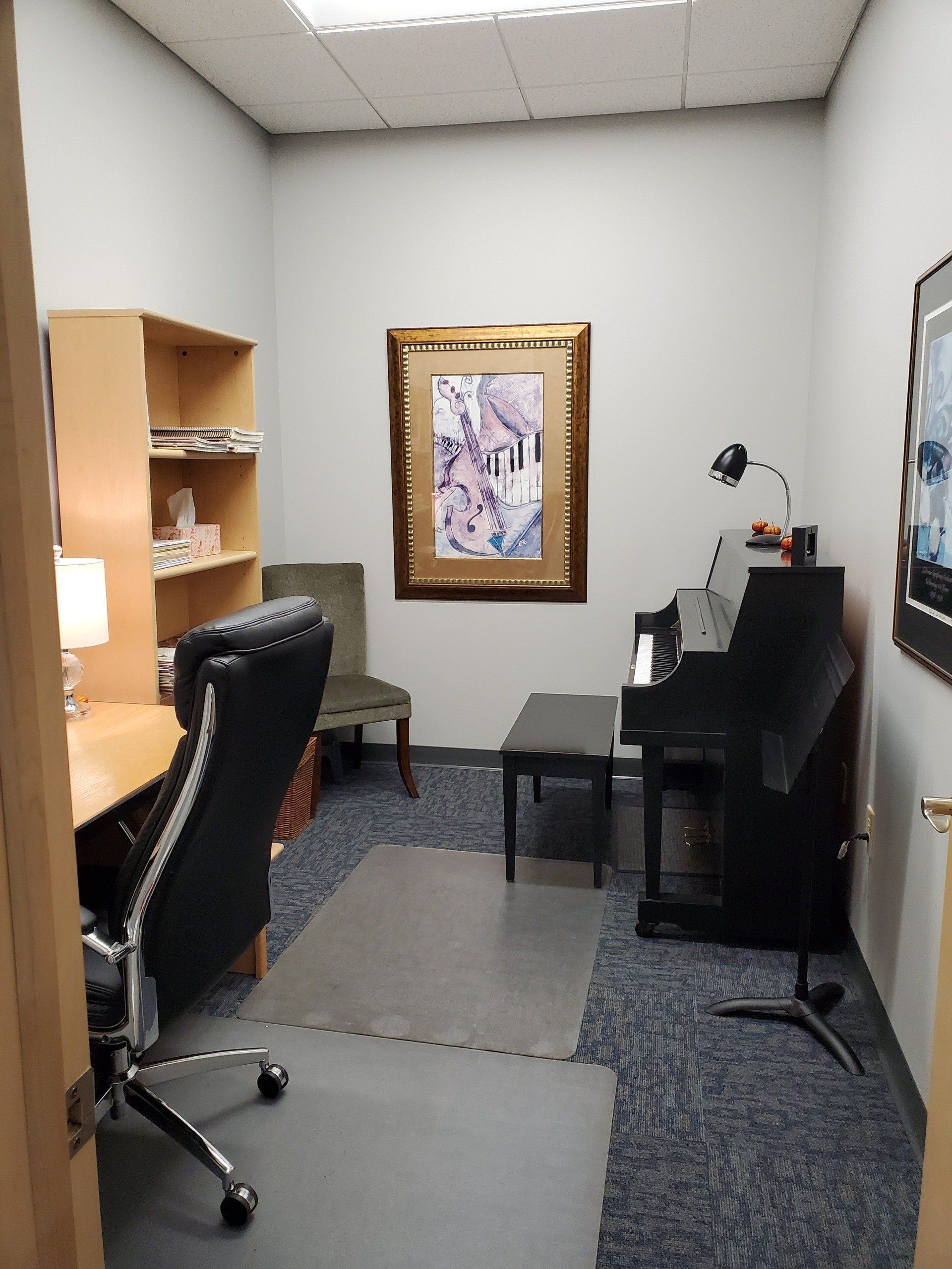 Small office with piano, desk, chair, and bookcase. Artwork on wall, gray carpet.