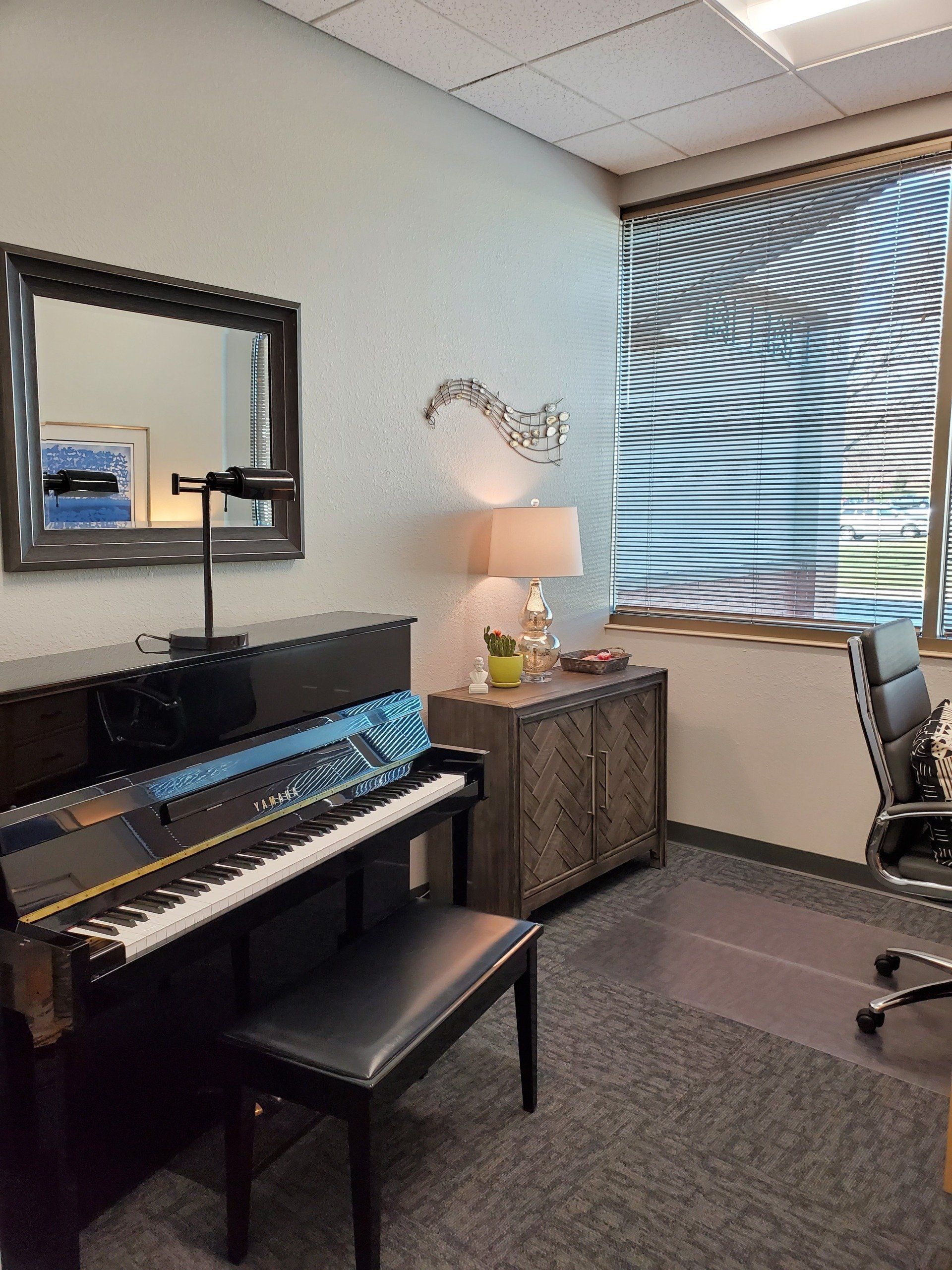 Piano in a room with a mirror, a small cabinet, and a window with blinds.