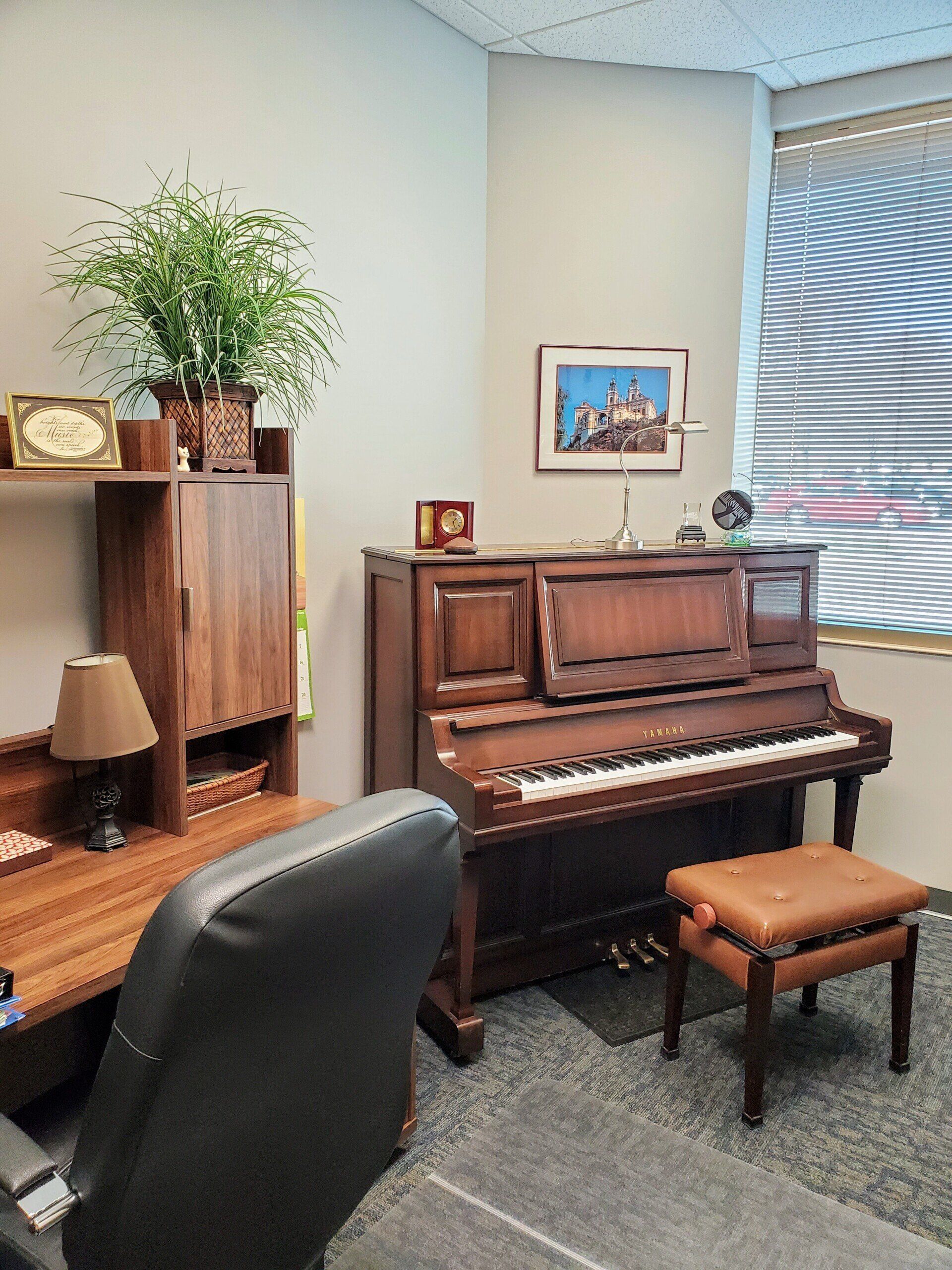 A piano and bench in a corner of an office, next to a desk and window with blinds.