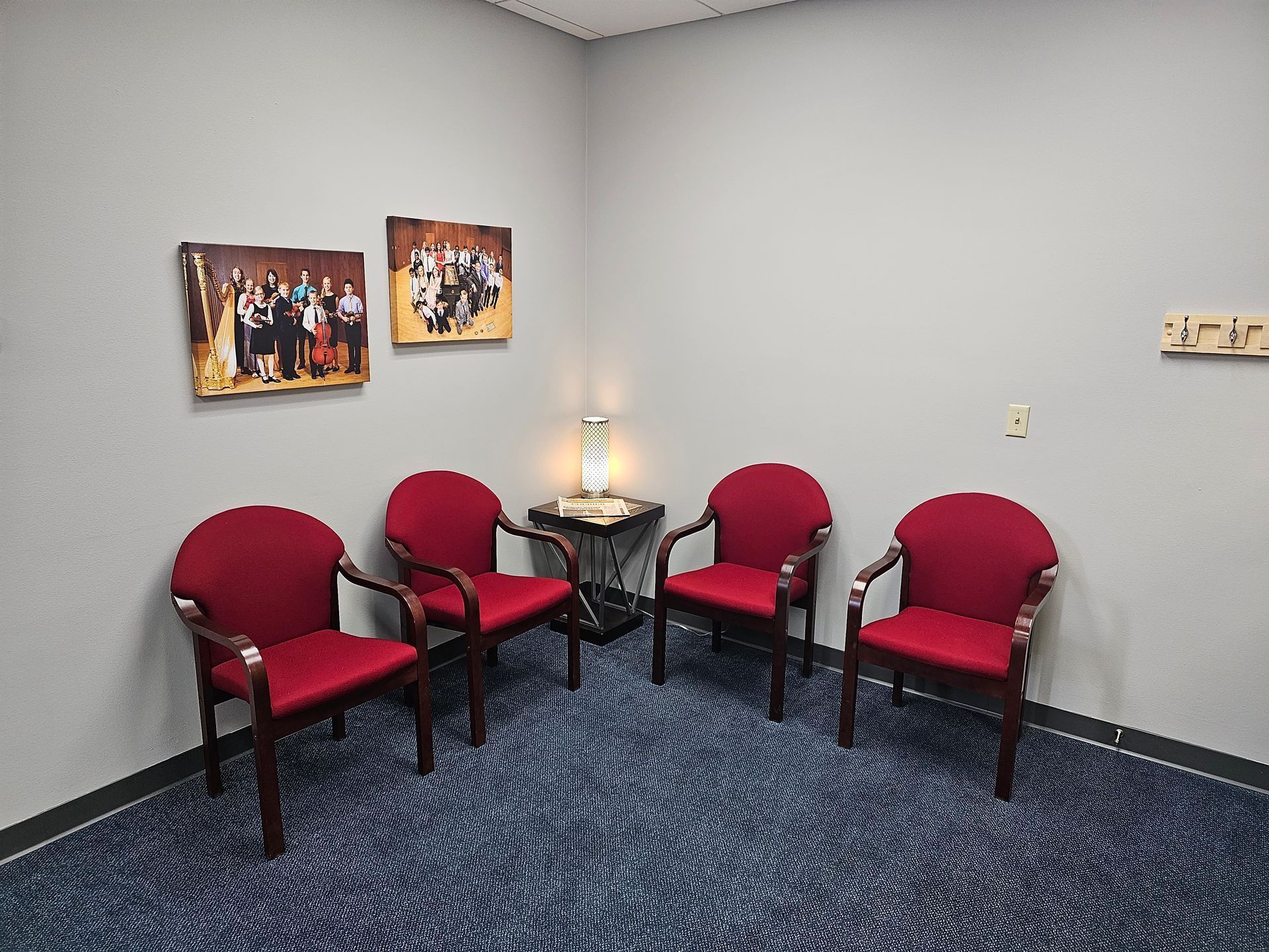 Four red chairs in a corner room with artwork and a small table with lamp. Blue patterned carpet.