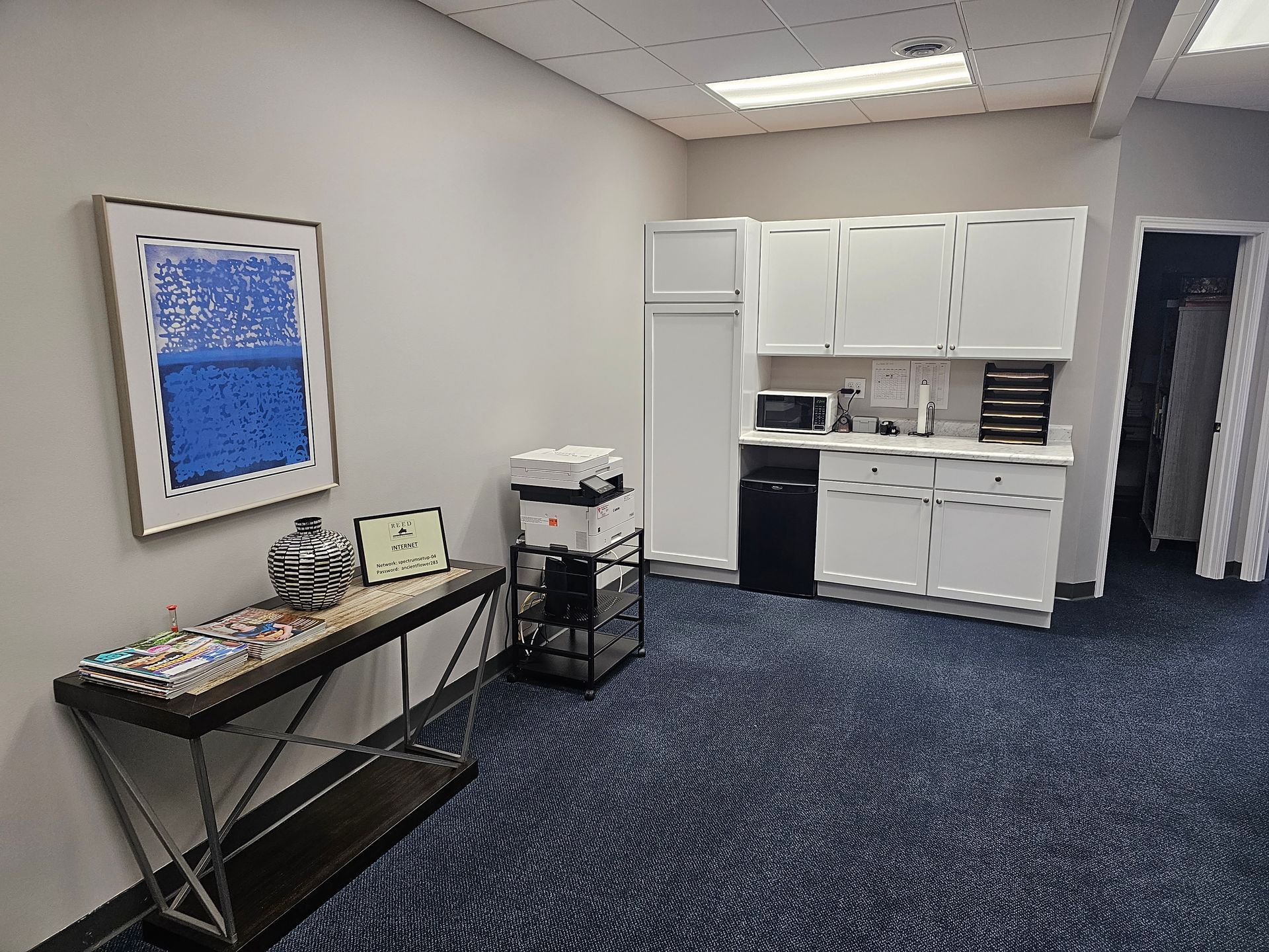 Office break room with a counter, cabinets, printer, and framed art. Blue carpet.