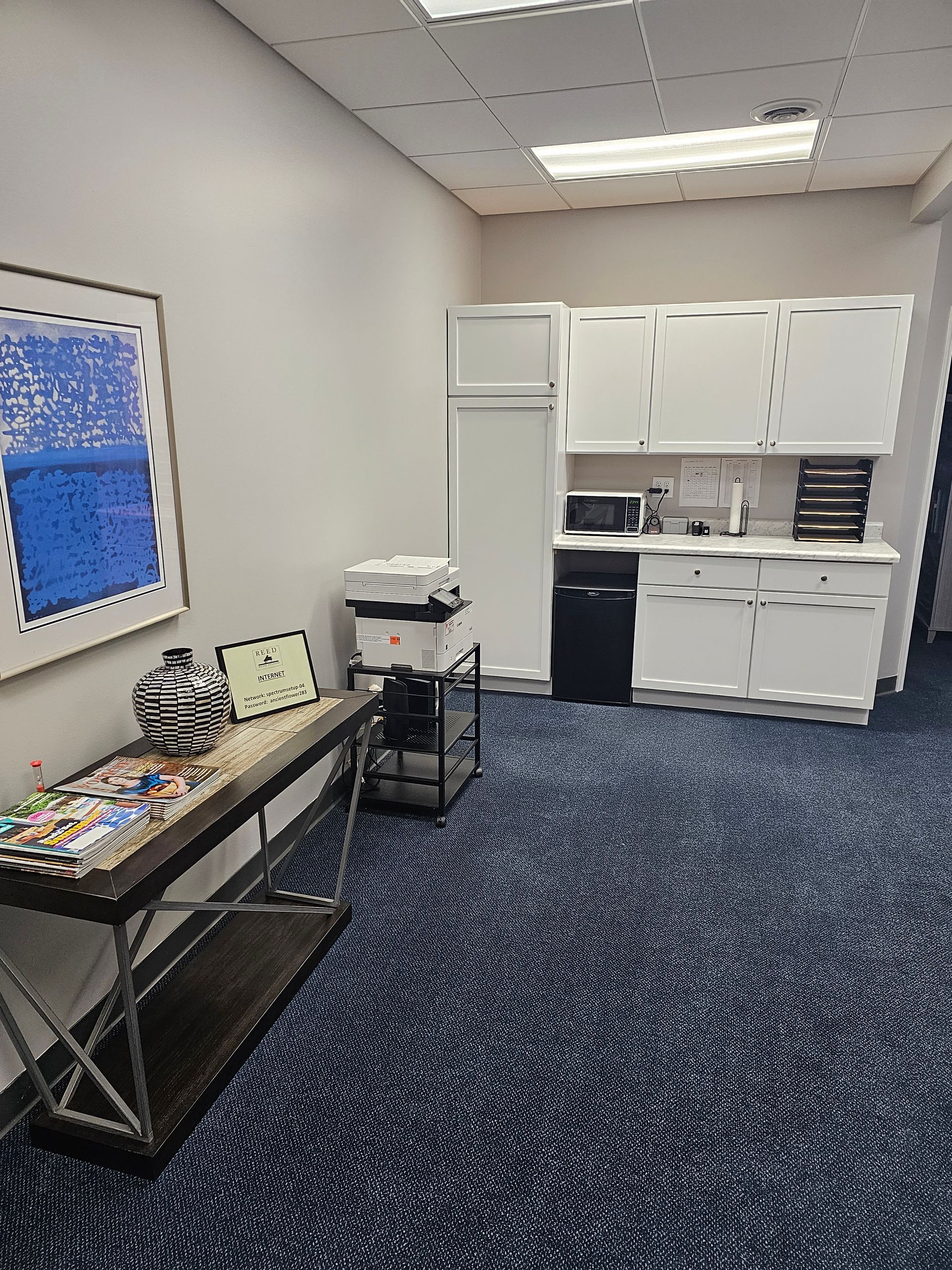 Office break room with a counter, cabinets, microwave, printer, and a table with art.