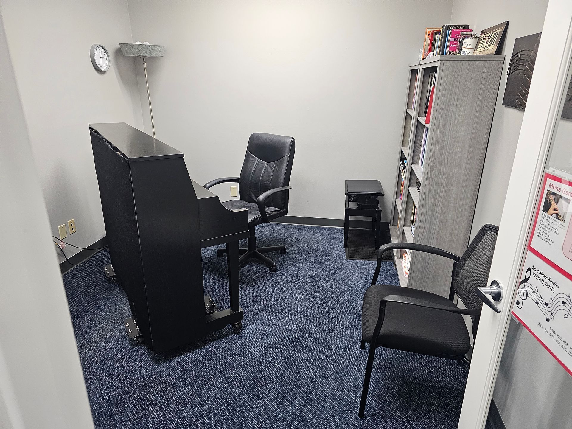 Piano room with black upright piano, chairs, bookshelf, and blue carpet.