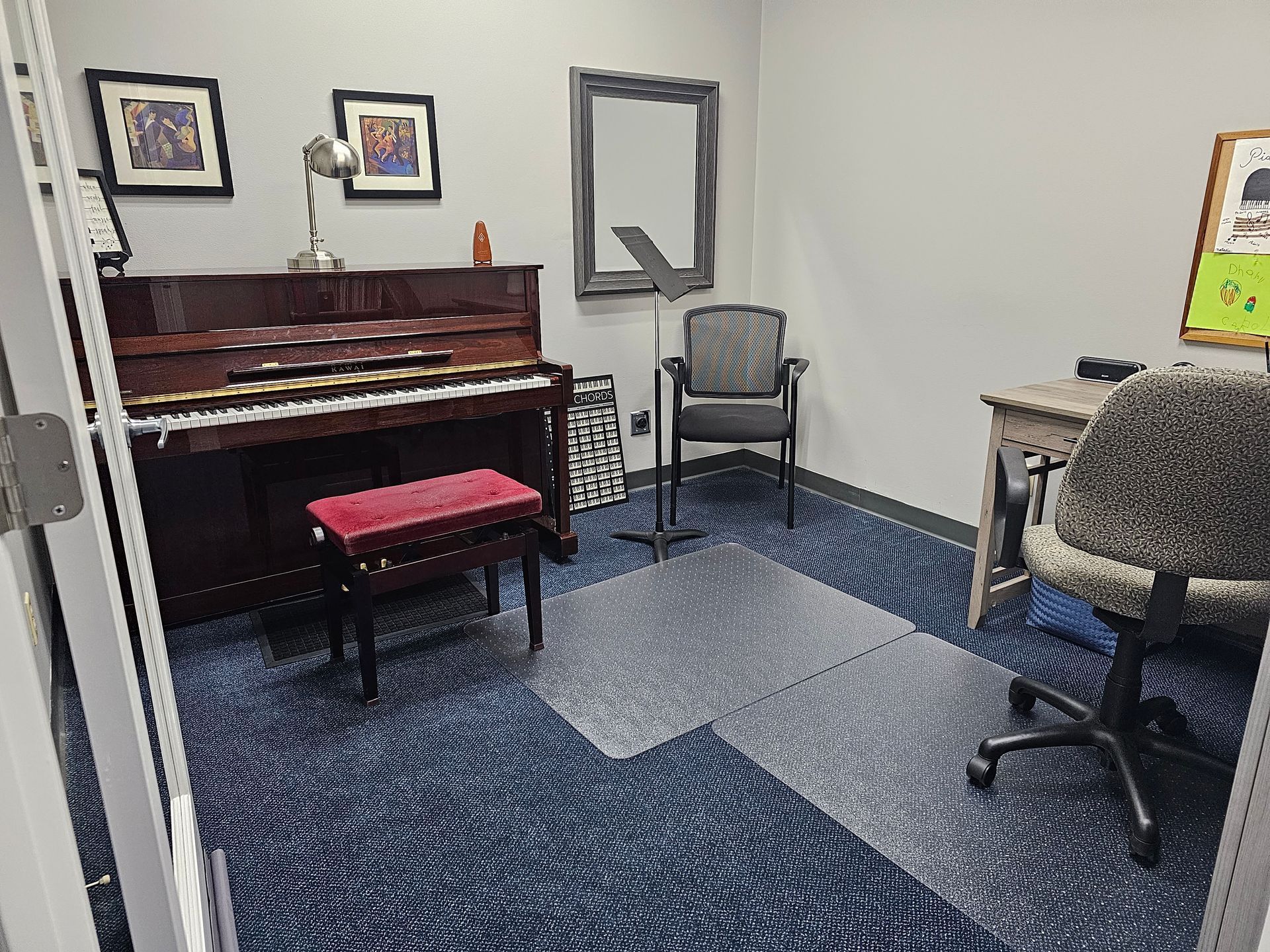 A music room with a piano, desk, chair, and sheet music. Dark-colored carpet and artwork on the walls.