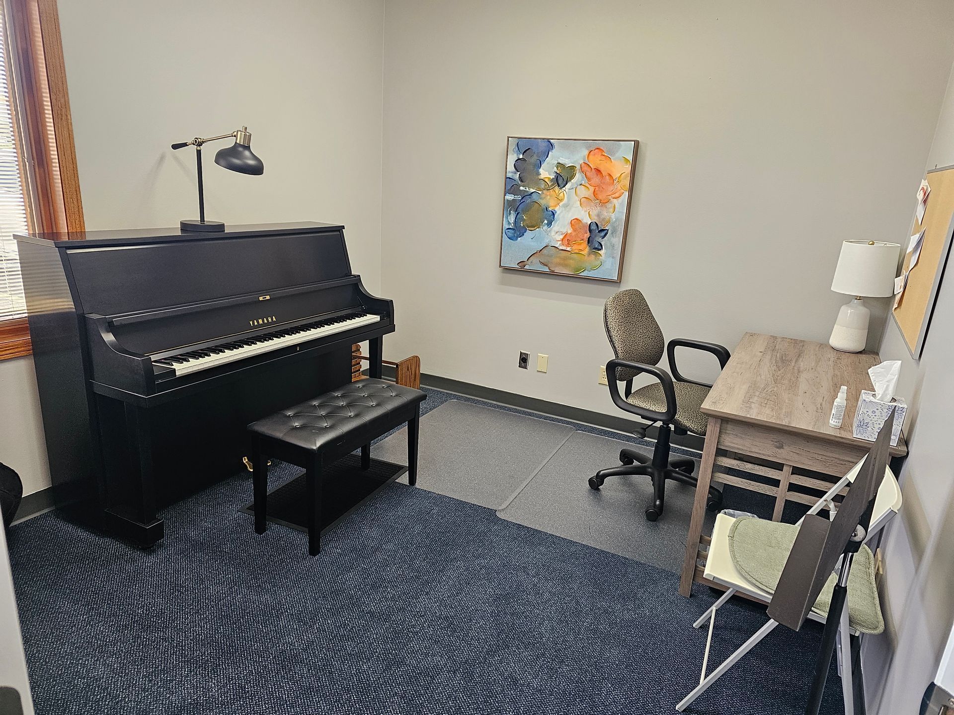 A room with a piano, desk, chair, artwork, and rug.