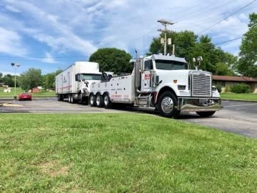 White tow truck towing a trailer on a grassy area. Blue sky and some trees are in the background.