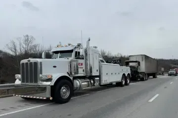 White tow truck towing a semi-truck trailer on a highway, overcast day.