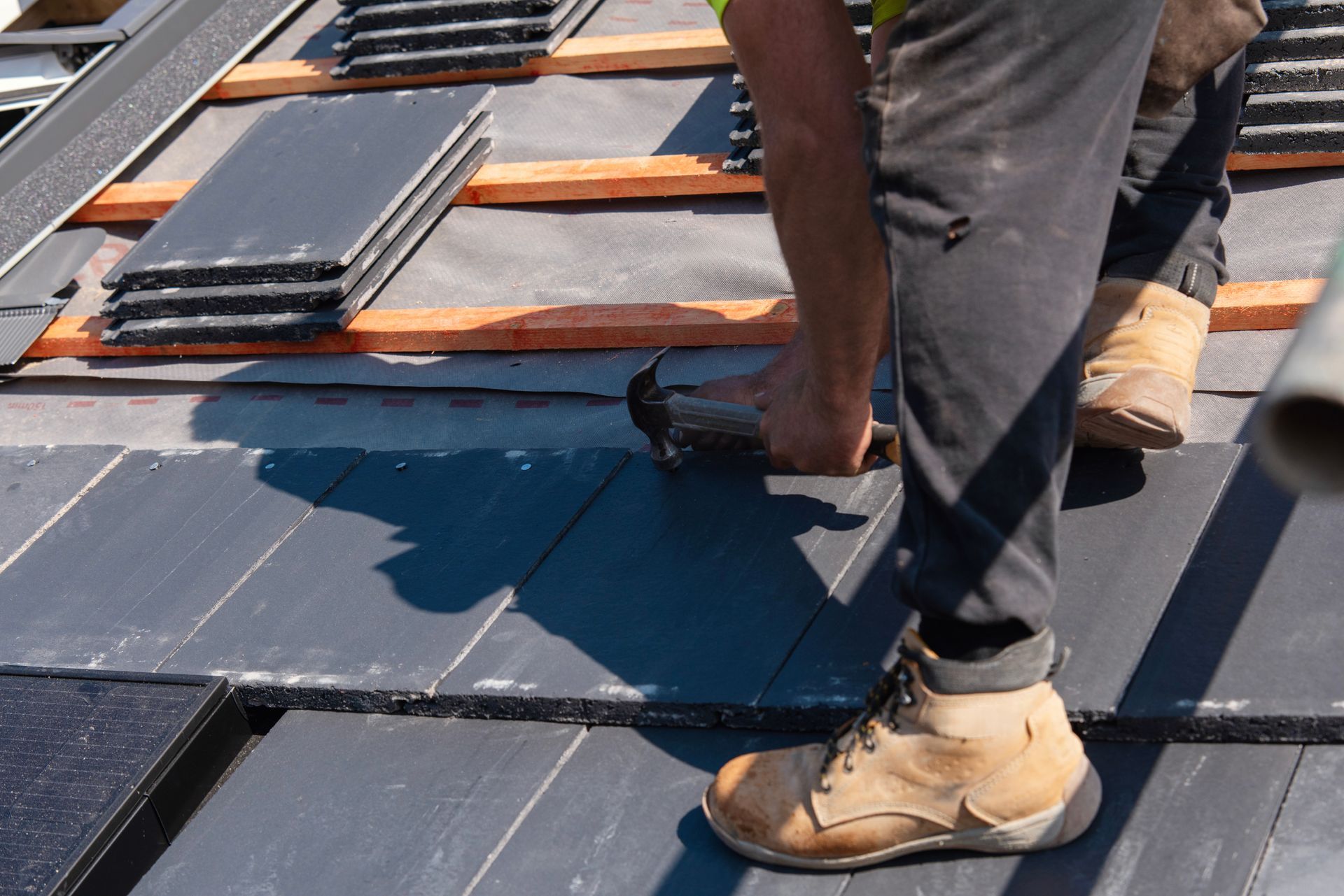 Worker installing dark roof tiles on a sloped roof with wooden battens visible