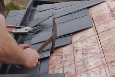 Worker installing dark roof shingles on a sloped roof, with exposed underlayment and tools visible.