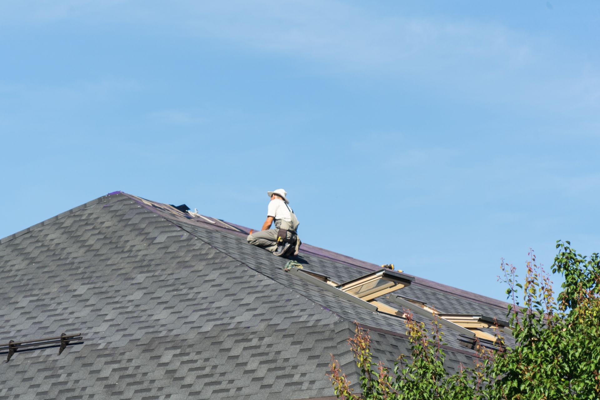 Roof worker repairing shingles on a gray roof under a clear blue sky