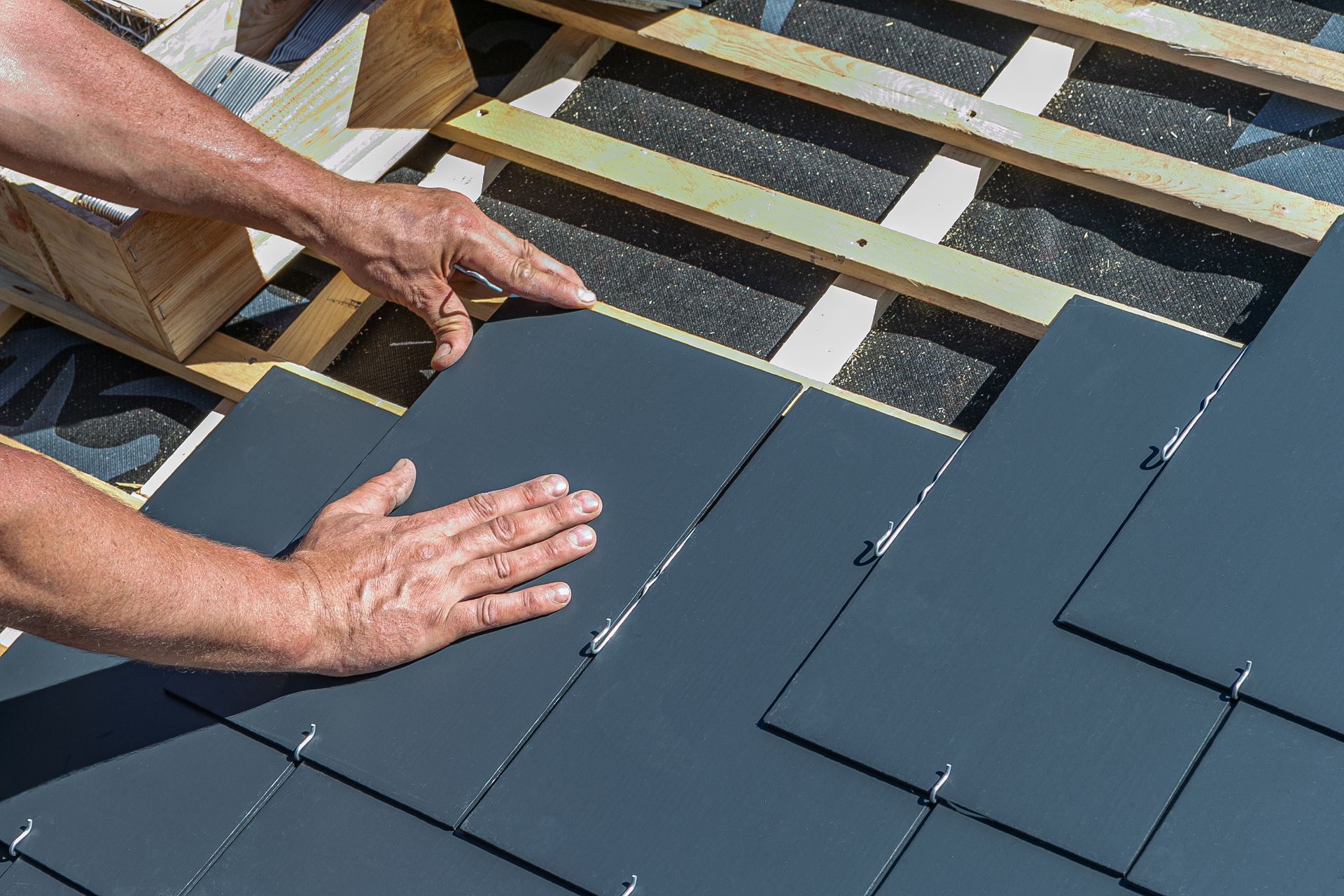 Hands installing dark gray roof shingles on exposed roof decking.