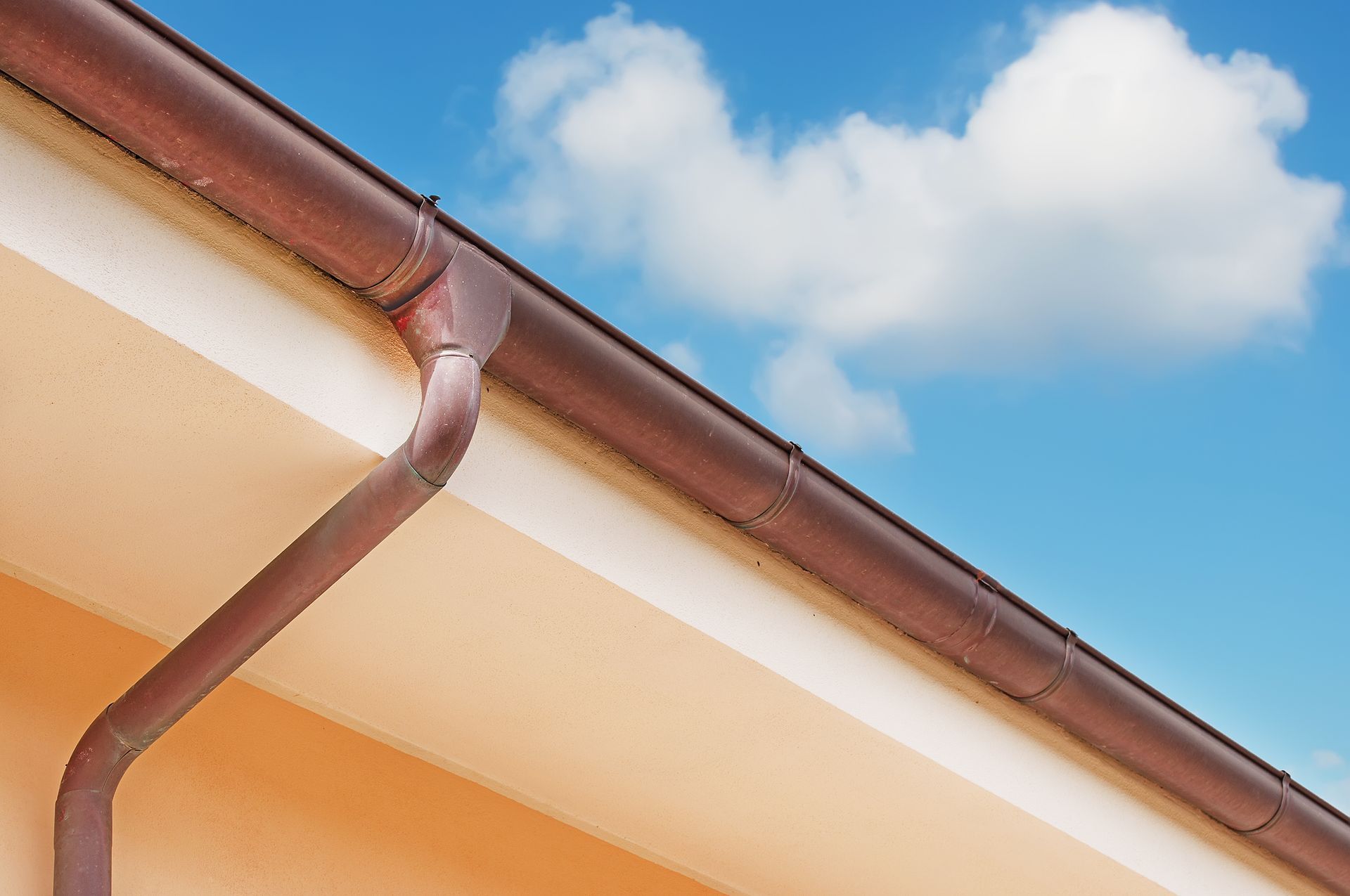 Brown rain gutter along a tan roof edge against a blue sky with clouds