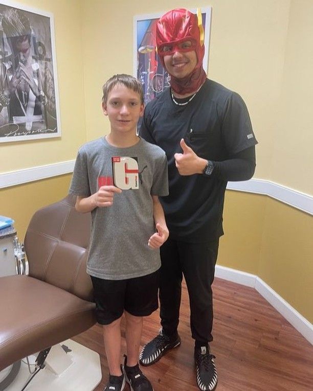 A boy wearing a gray shirt with the letter g on it stands next to a man wearing a red mask