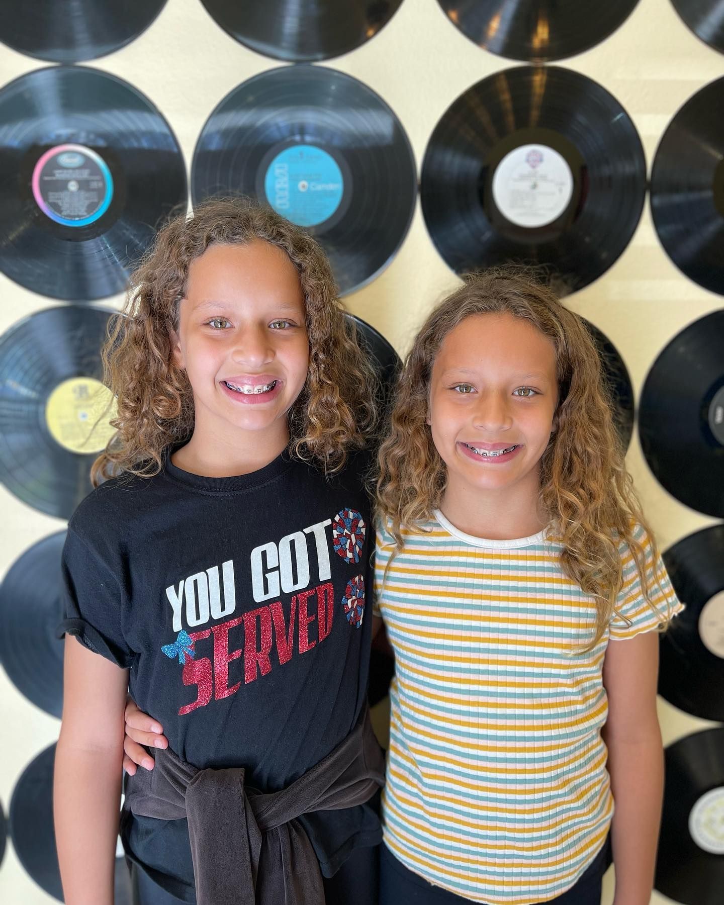 Two young girls are posing for a picture in front of a wall of vinyl records.