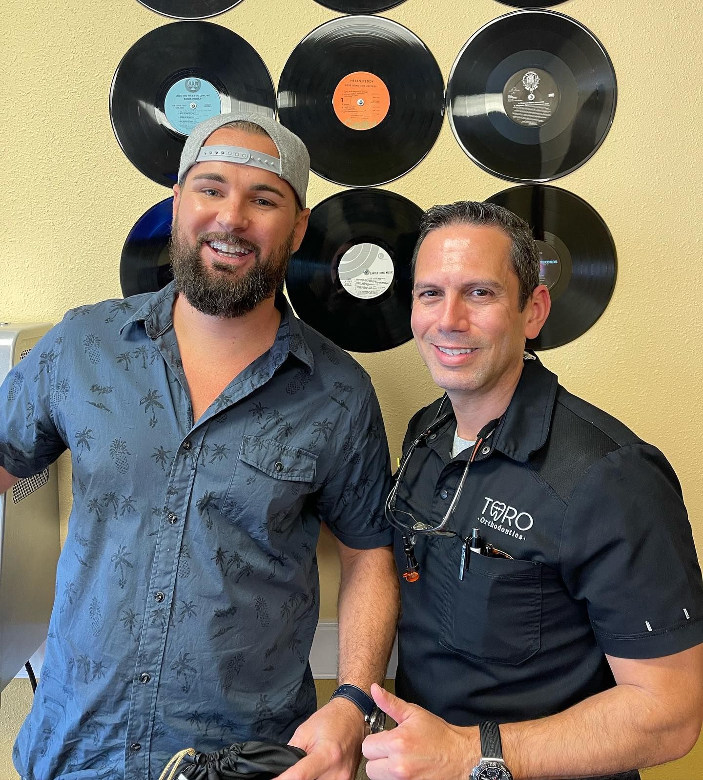Two men are posing for a picture in front of a wall of vinyl records