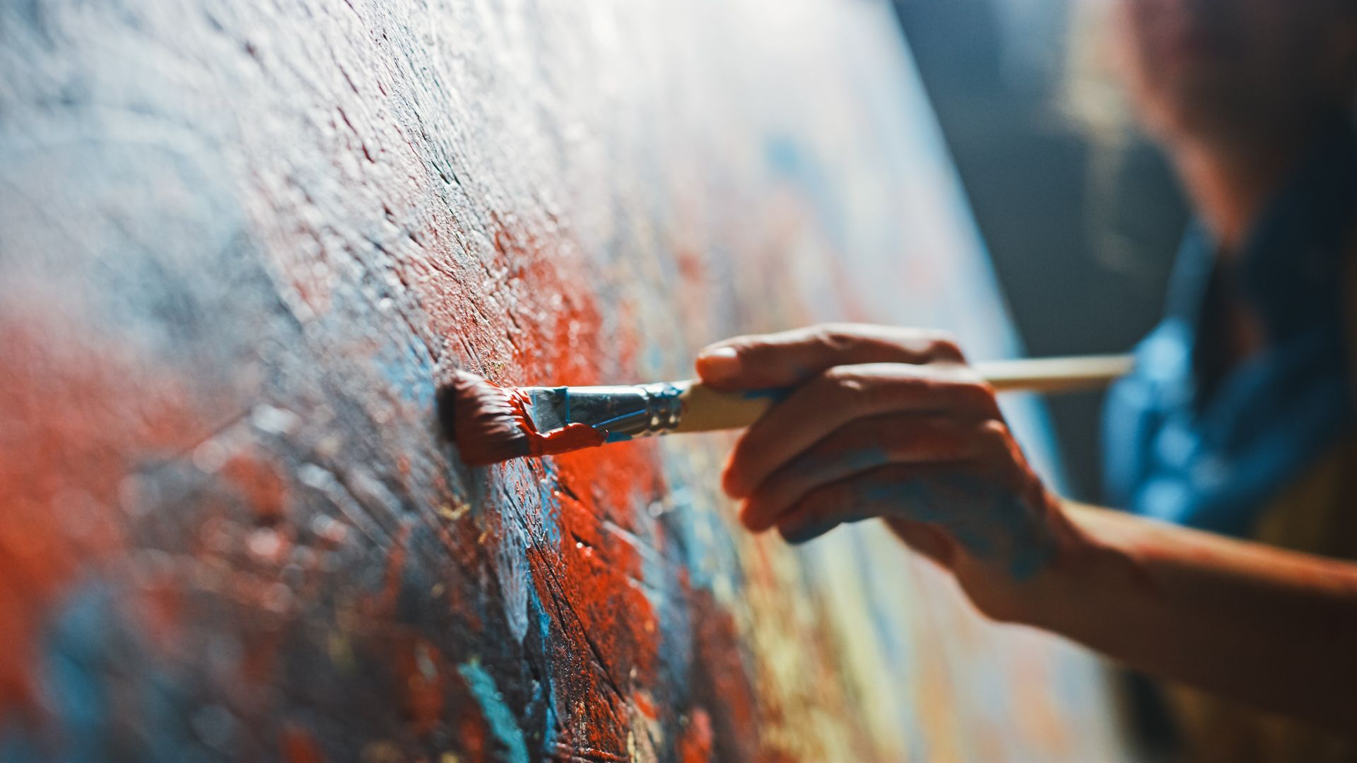 A close-up of a hand holding a paintbrush, applying textured red, orange, and blue paint to a canvas.