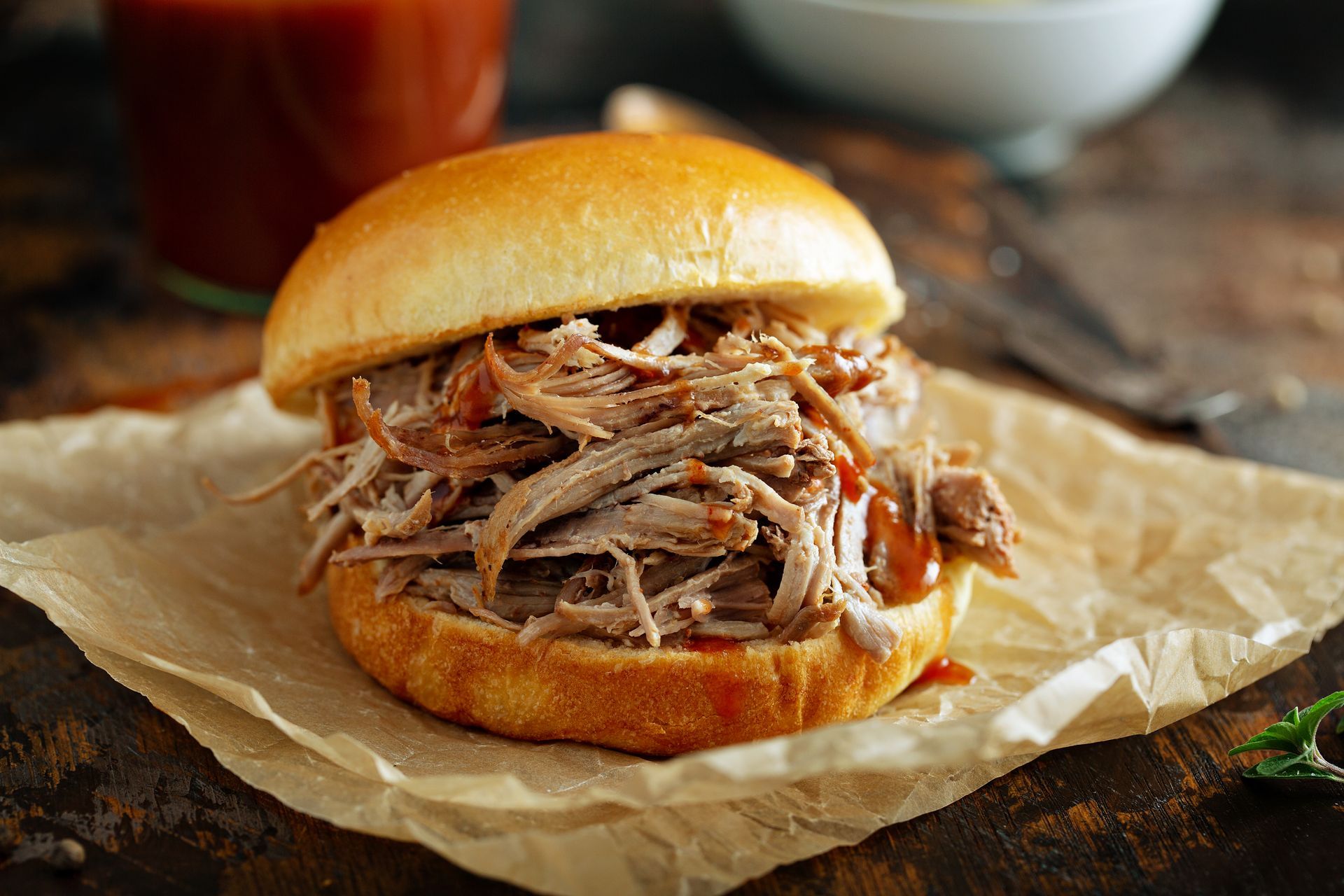 A pulled pork sandwich with barbecue sauce on a toasted bun, resting on parchment paper on a rustic wooden surface.