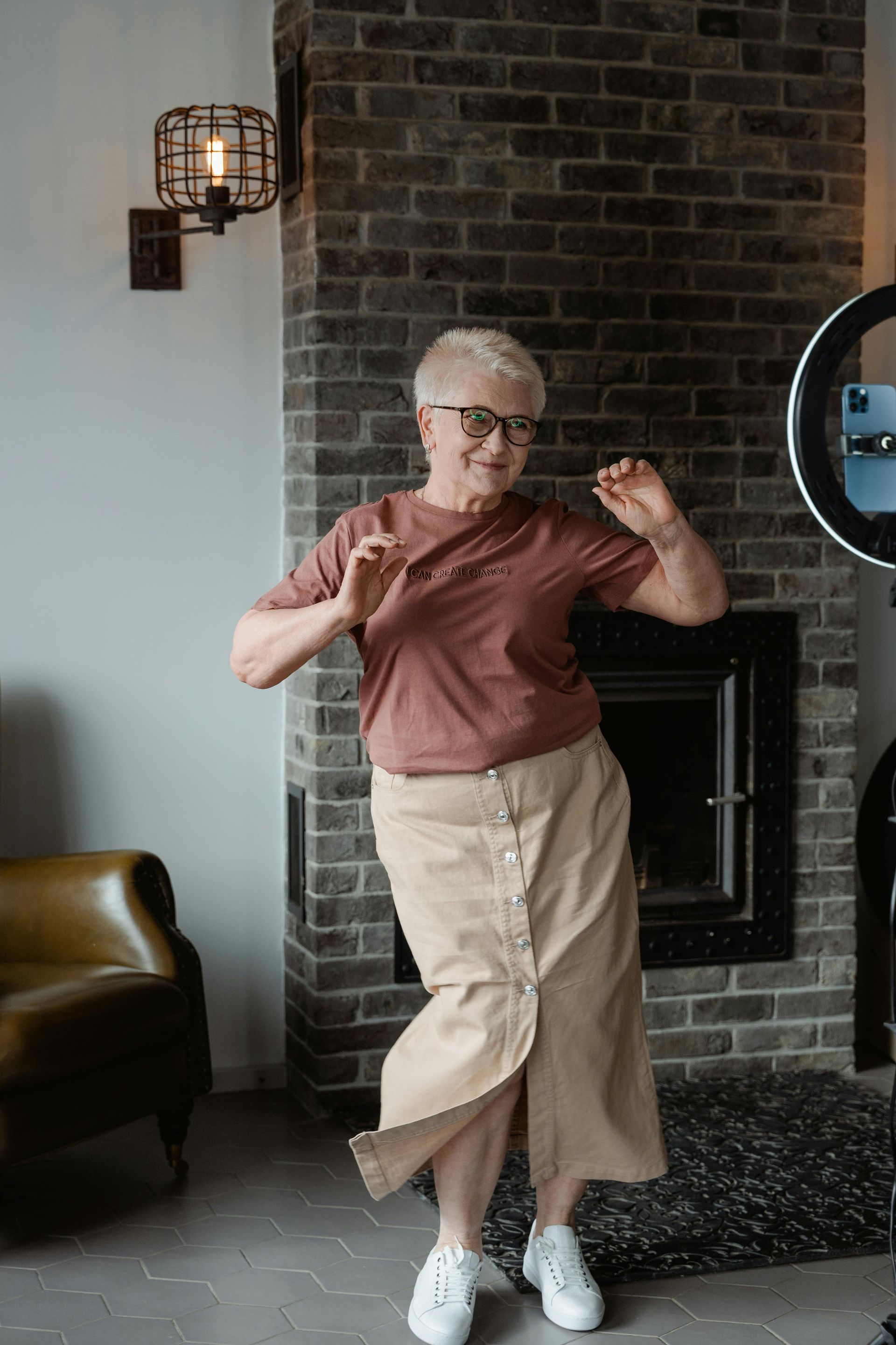 An elderly woman is dancing in front of a brick fireplace in a living room.