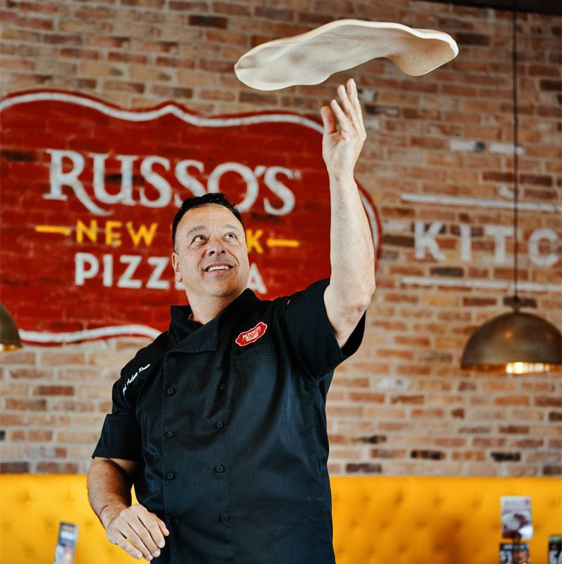 Chef tossing pizza dough, smiling in a Russo's New York Pizza Kitchen. Brick wall, interior.