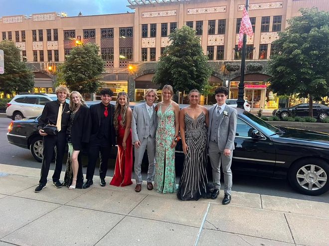 Group posing on a sidewalk in formal evening wear in front of parked cars and a lit building.