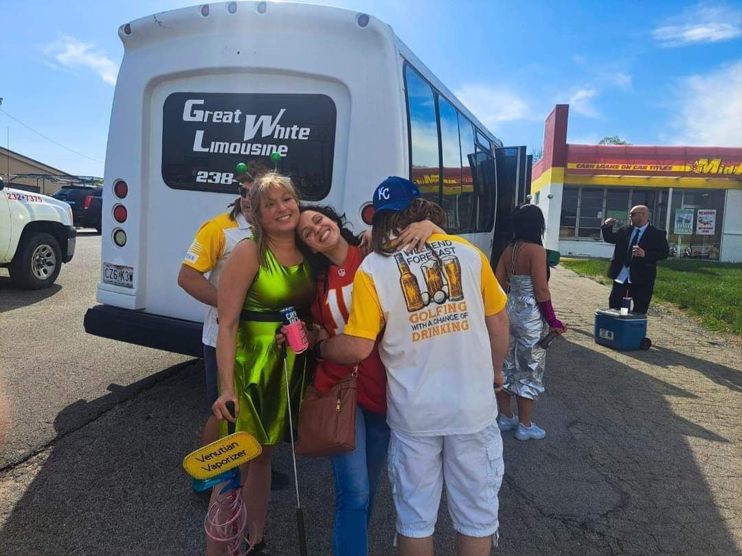 People posing beside a white tour bus in a parking lot, with a storefront in the background