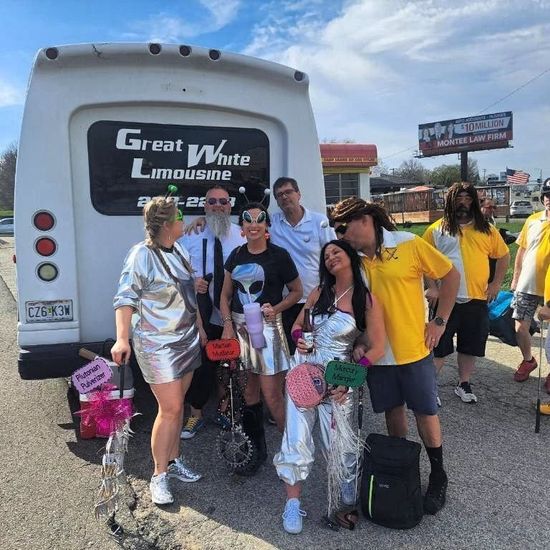 Group posing in front of a Great White Limousine van at an outdoor event, with bags and props.