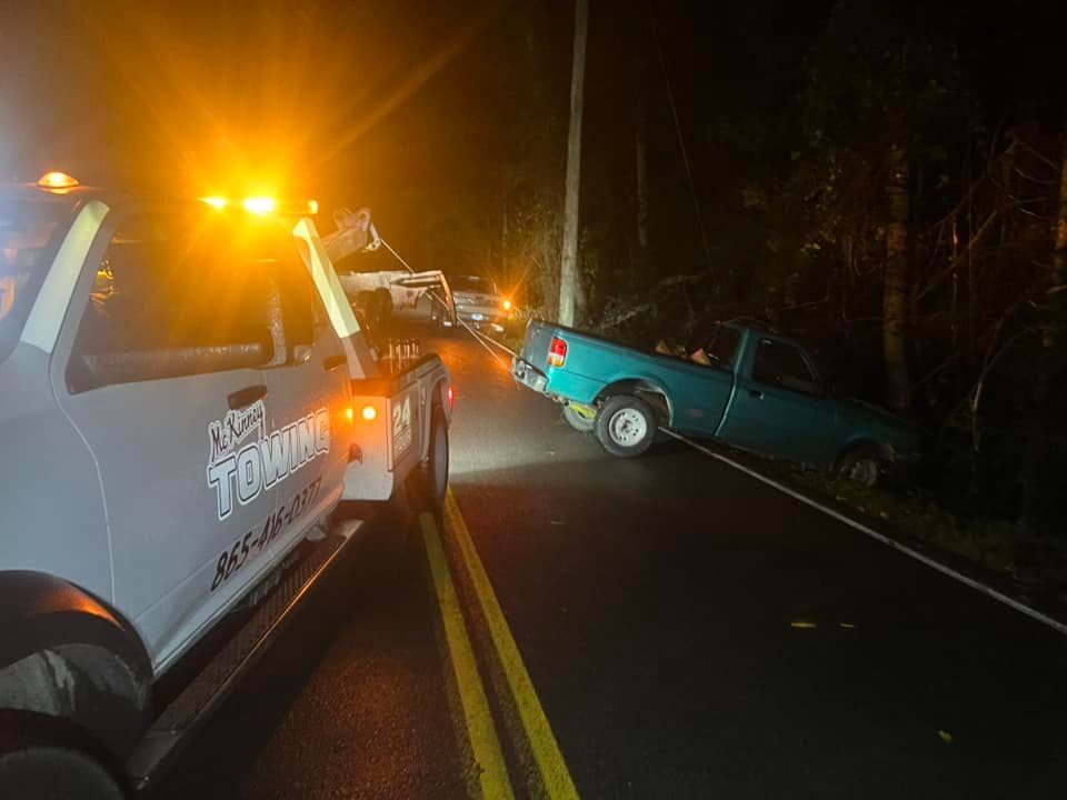 A tow truck is towing a blue truck down a road at night.