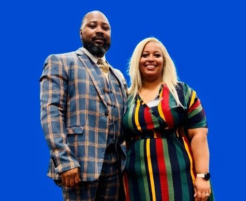 A smiling man in a plaid suit and a woman in a stipe dress standing against a solid blue back ground.