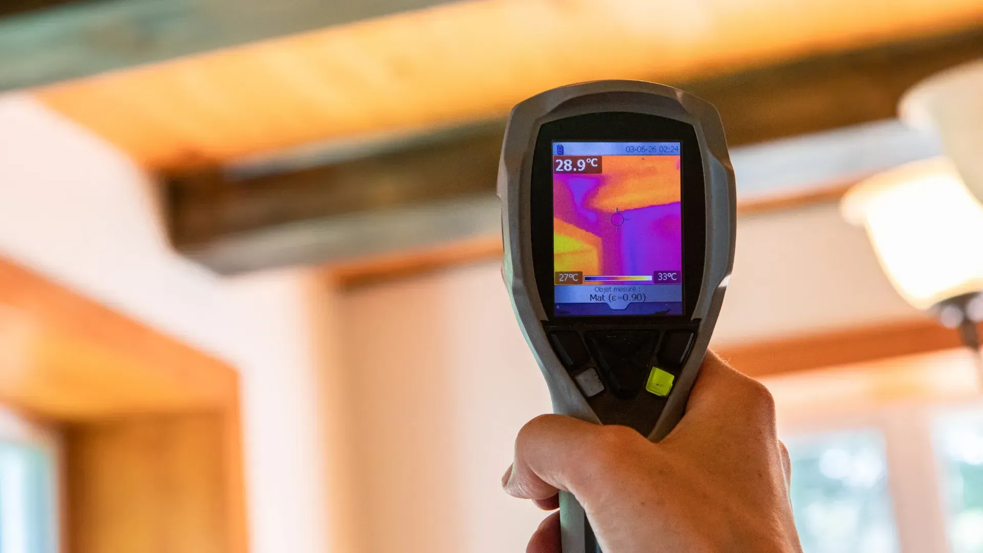 Hand holding a thermal imaging camera, pointing it at a beam, displaying heat variations with color on the screen.