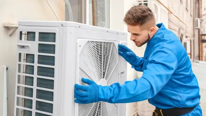 Man in blue jumpsuit inspecting a white outdoor air conditioning unit, wearing gloves.