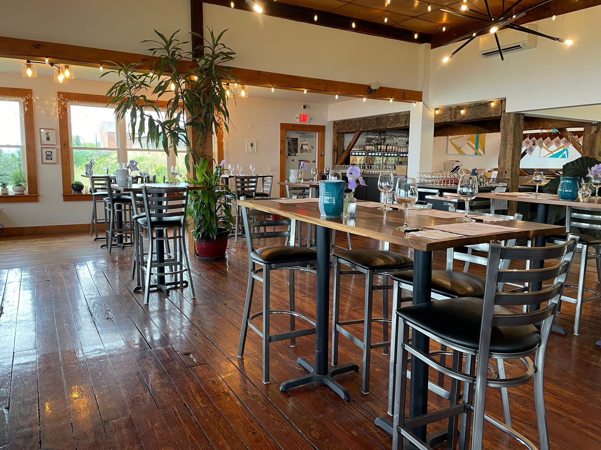 An open-plan dining room with high wooden tables, black metal bar stools, and polished wooden floors under warm lighting.