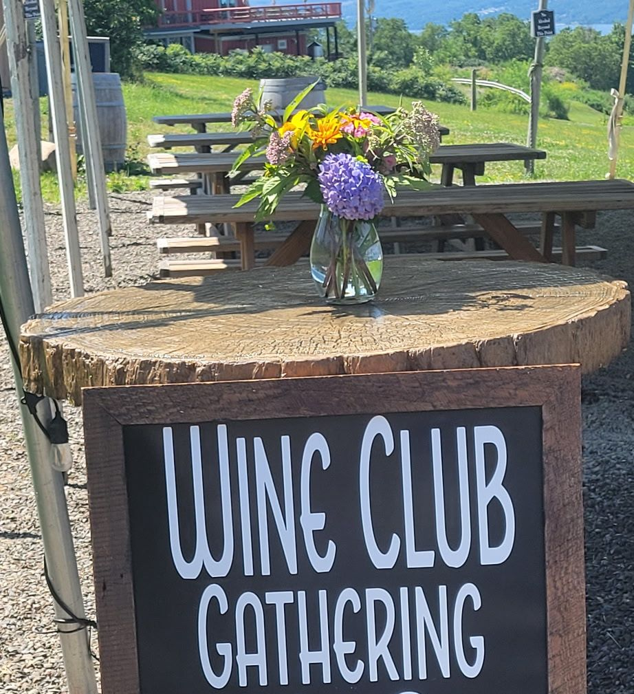 Wine club gathering sign beside a picnic table with a flower arrangement outdoors.