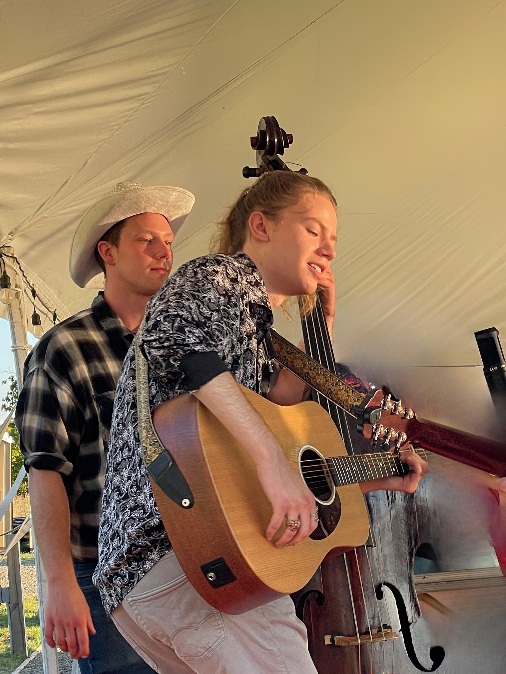 Two musicians performing indoors under a white tent, one singing and playing acoustic guitar.