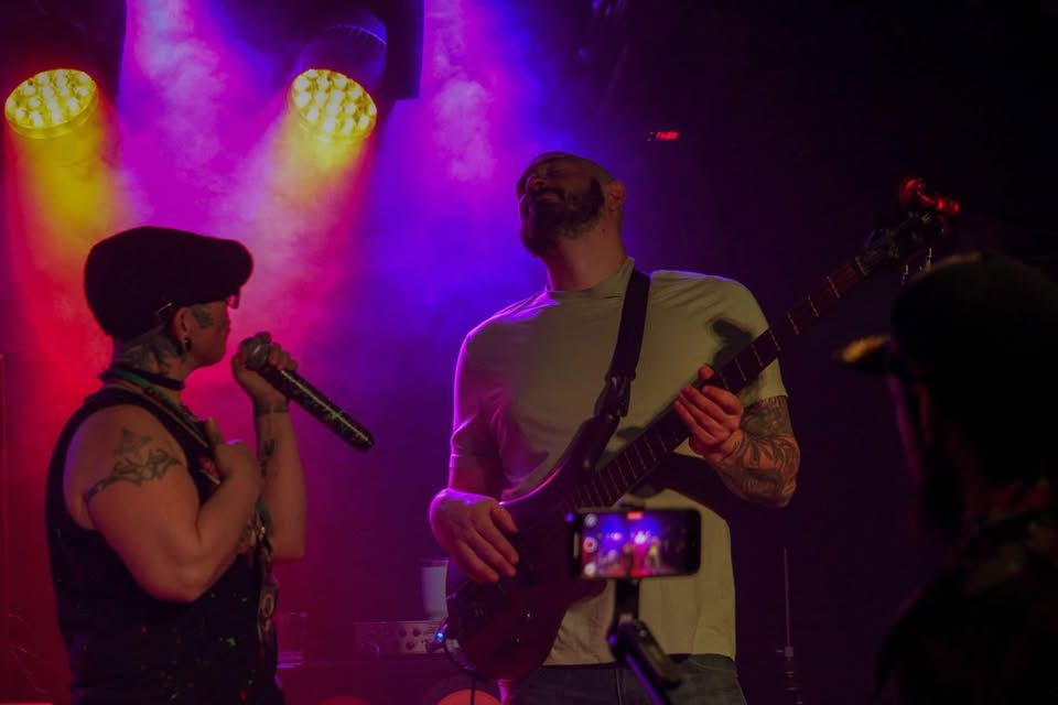 Two musicians performing on a purple-lit stage, one singing into a microphone and one playing bass.
