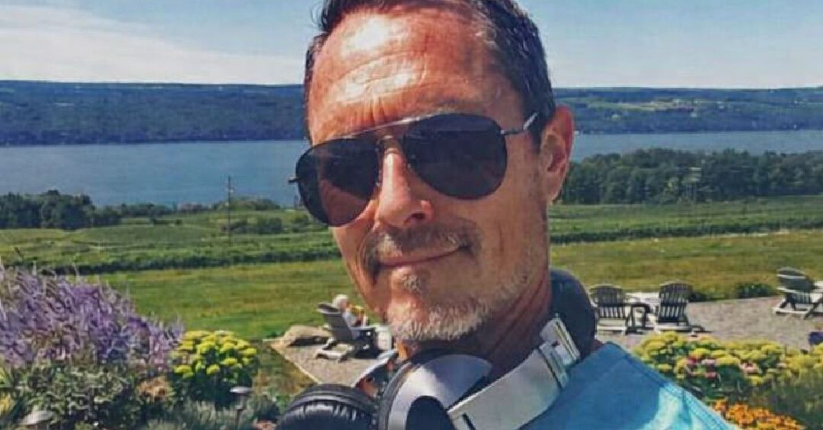 Man wearing sunglasses and headphones takes a selfie with a lake and green hills in the background