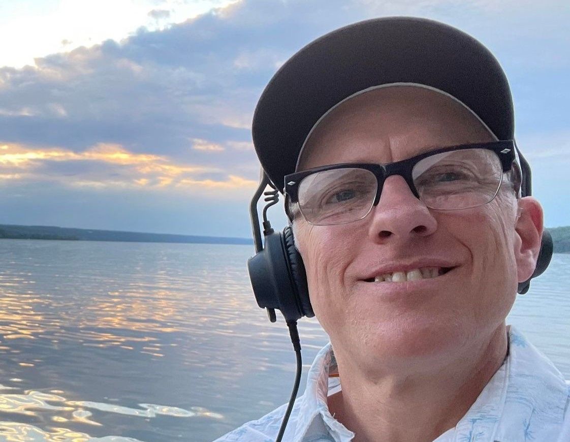 Smiling person wearing glasses and a cap by a calm waterfront at sunset