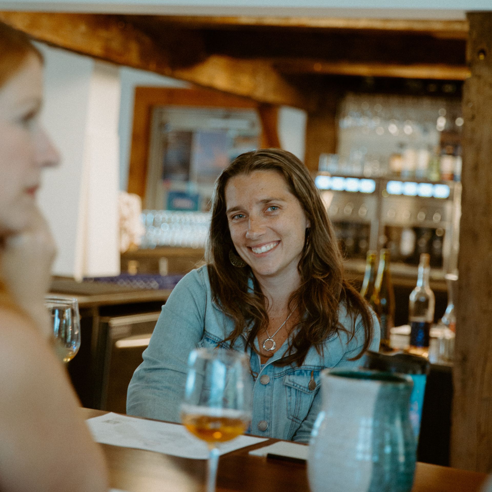 A smiling person in a denim jacket sits at a table with a wine glass in a rustic, dimly lit restaurant setting.