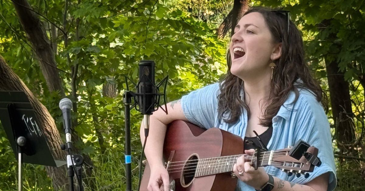 Woman singing while playing acoustic guitar outdoors in a green park setting