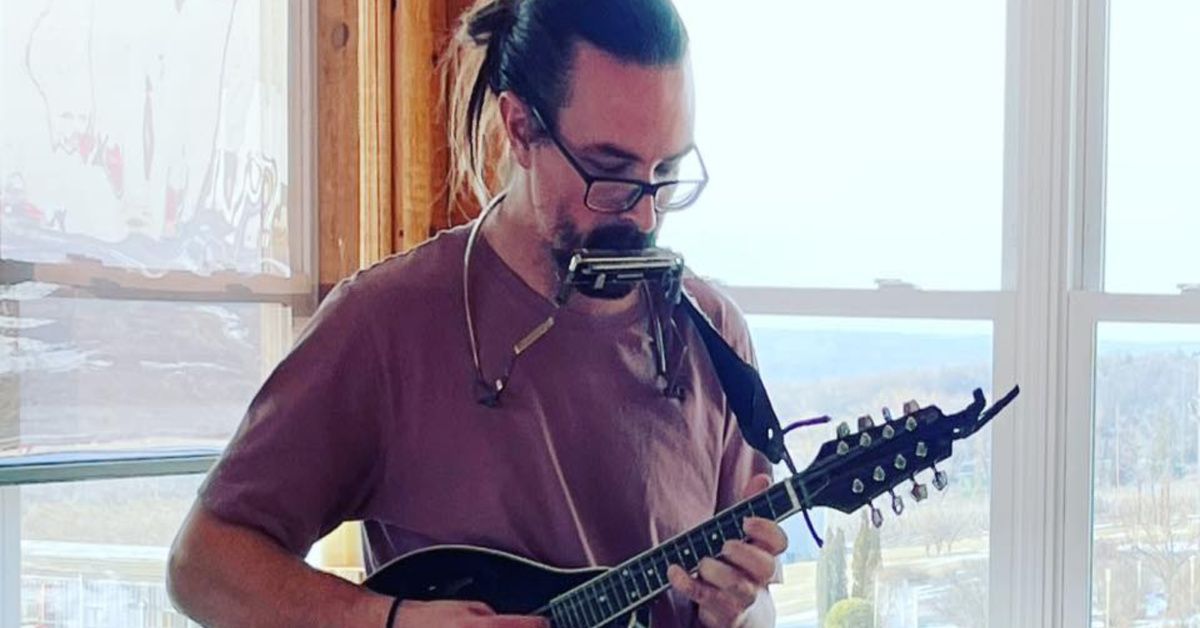 Man playing mandolin by a window, wearing glasses and a maroon shirt, with a harmonica holder around his neck